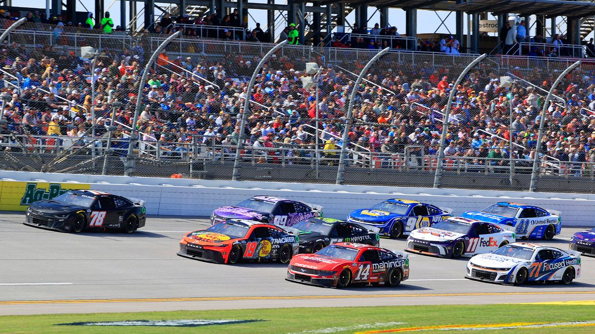 A telephoto shot of NASCAR cars driving along Talladega Superspeedway, with the crowd visible behind a large barrier in the background.