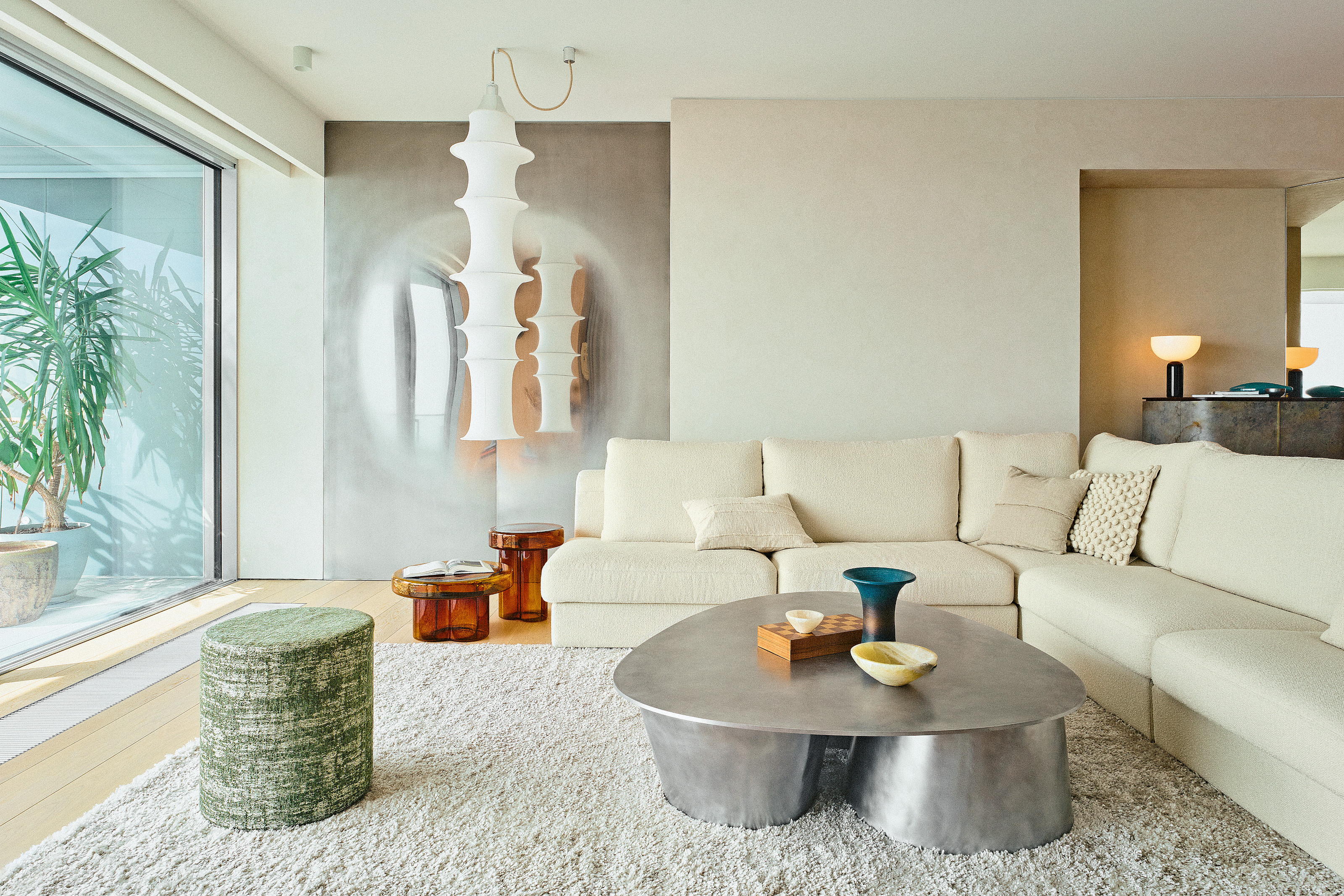 Living room with cream textured walls, cream corner sofa, aluminium coffee table, green stool, amber glass side tables and white paper pendant lights