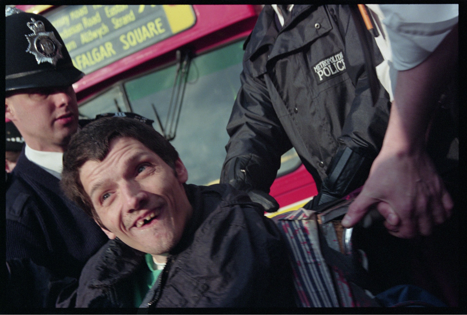 A wheelchair user in a pink sweatshirt holds a sign reading "How about us?" beside a red London Leyland bus, as bystanders look on from the pavement.