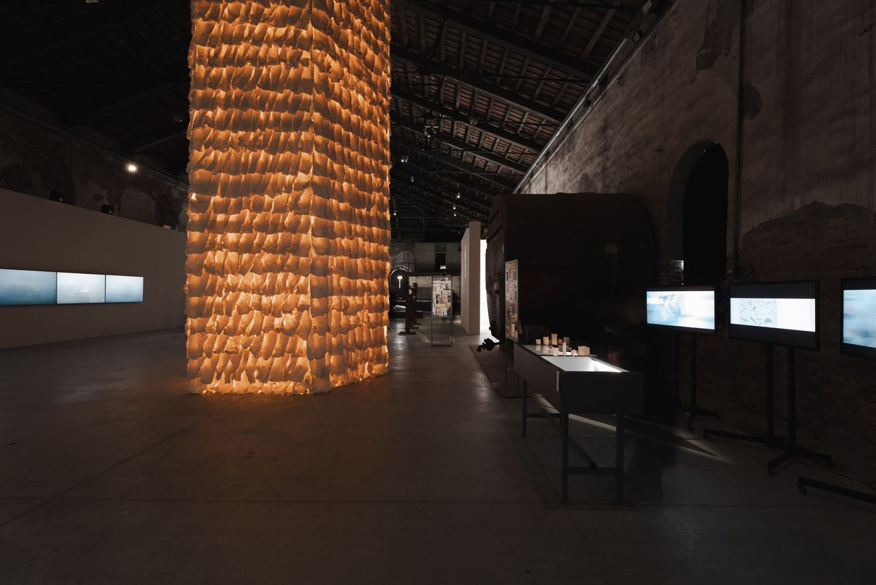 A towering, softly glowing sculpture made of paper sits at the center of a darkly lit, brutalist gallery.