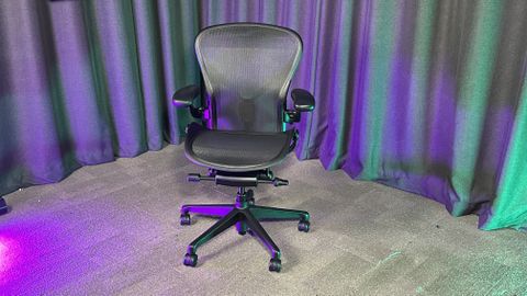 The Herman Miller Aeron gaming chair on a grey floor with grey curtains behind it