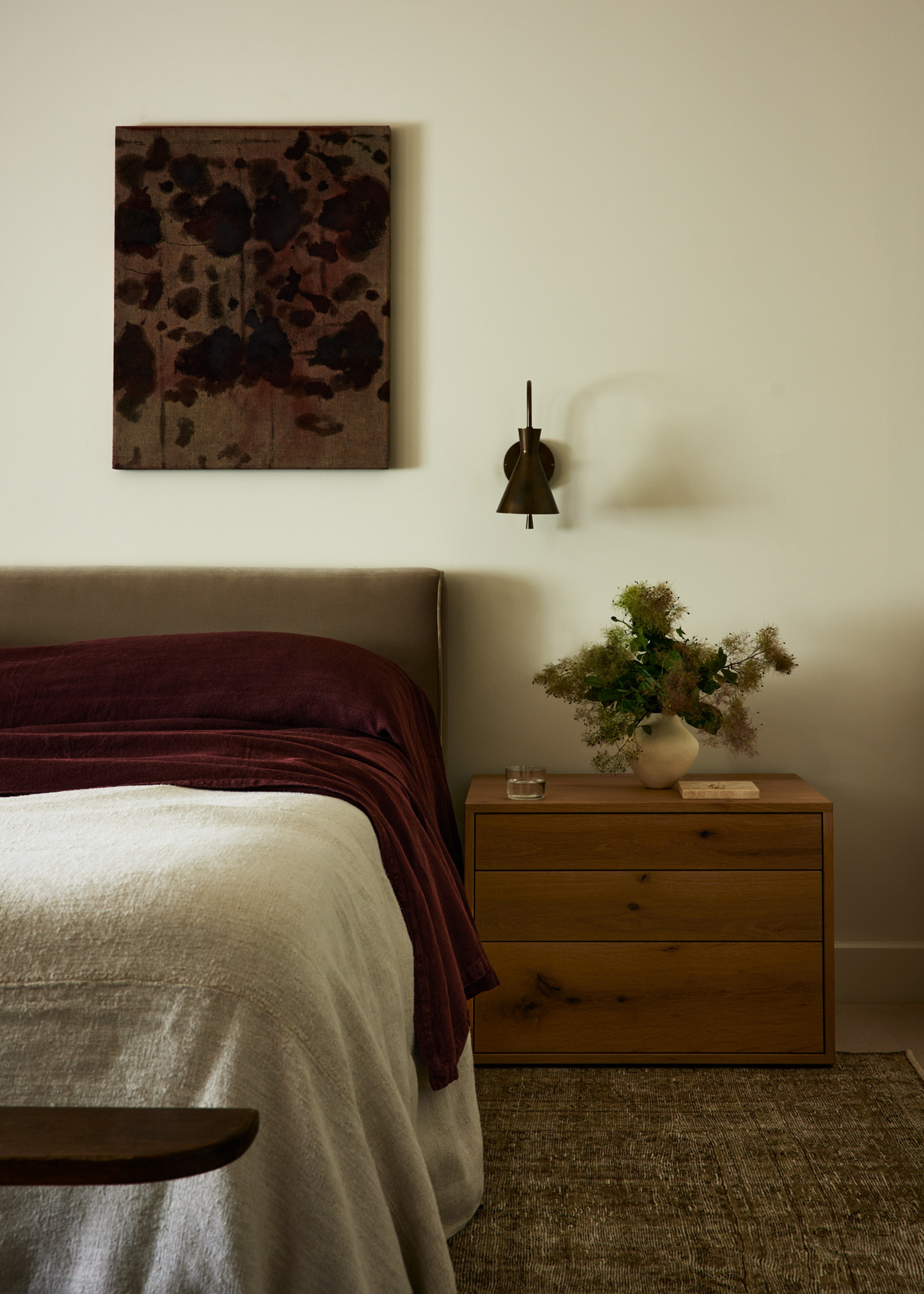 modern bedroom with timber side table, bed with upholstered velvet headboard, vintage rug, wall sconce and off-centre artwork