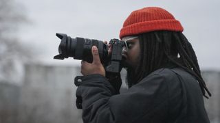 Screenshot from Adorama TV episode of Picture America of photographer Pat Crey, side-on, wearing an orange beanie hat and looking through the viewfinder of a Sony camera