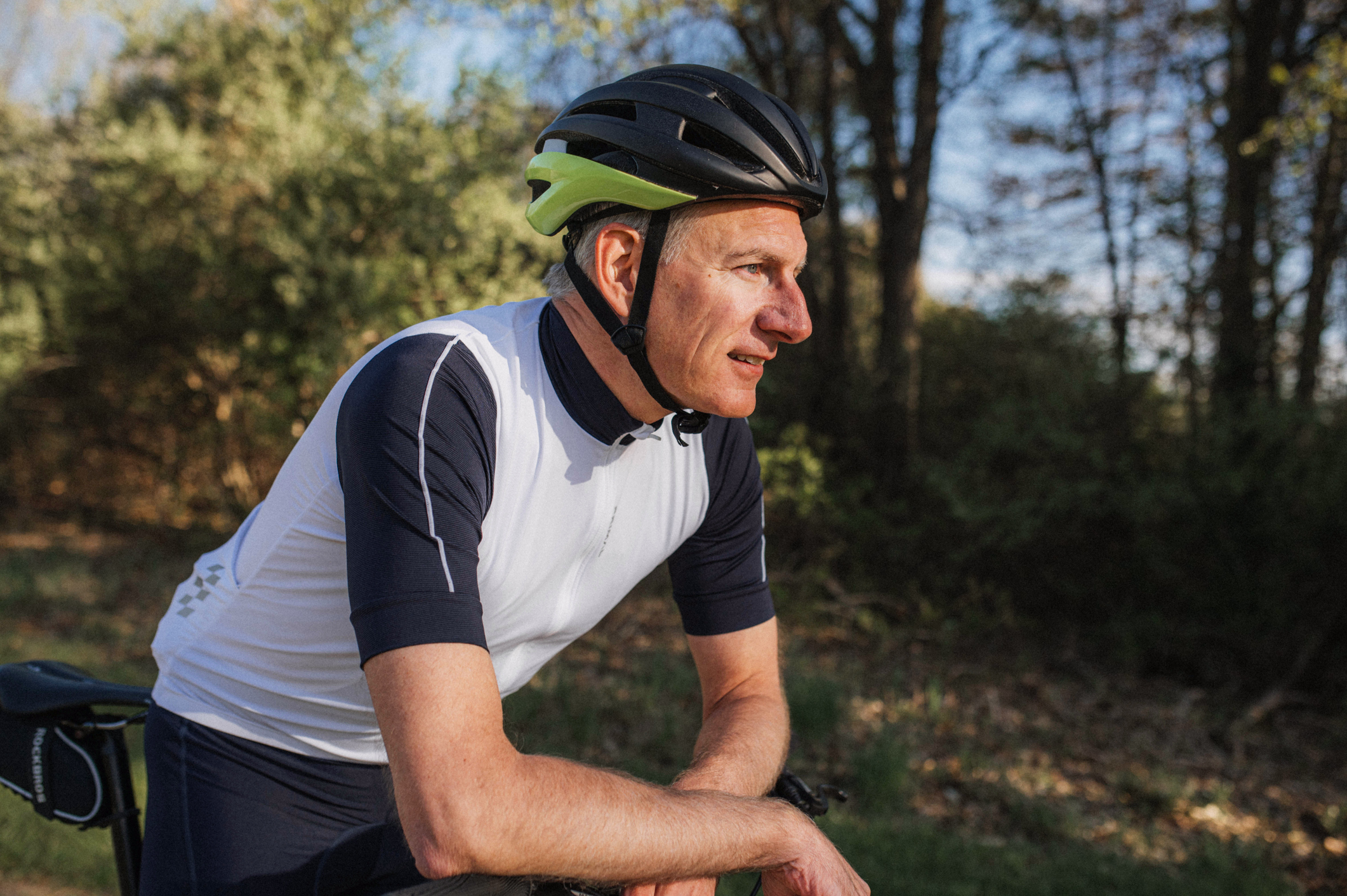 A cyclist in a helmet rests by his bicycle in a wooded area. The sunlight filters through the trees, creating a serene atmosphere.