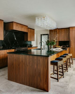 kitchen island with dark wood cabinets black countertops and backsplash and pale floor tiles with intricate clear glass light fitting above island