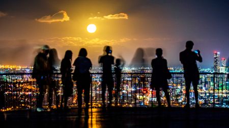 Silhouettes of people standing on a balcony overlook the cityscape of lights below with a full glowing moon in the distance.