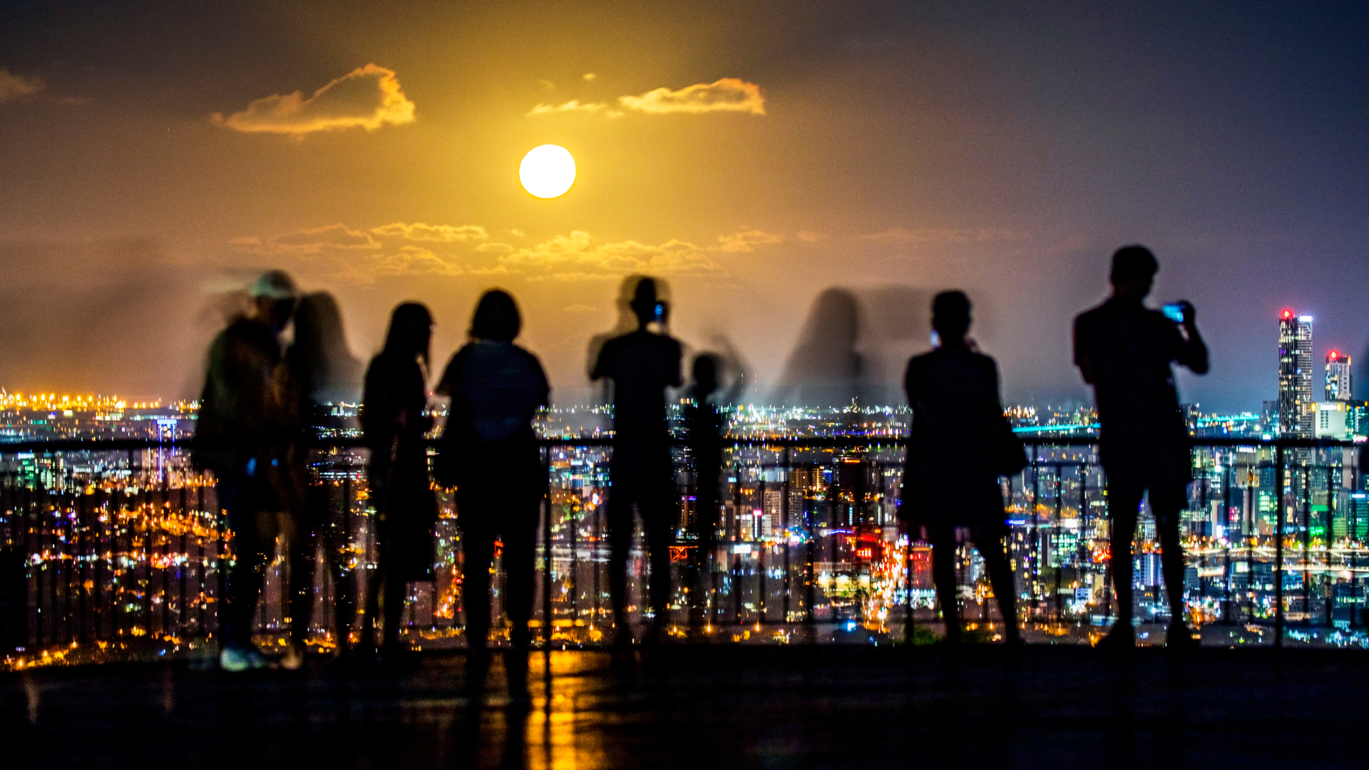 Silhouettes of people standing on a balcony overlook the cityscape of lights below with a full glowing moon in the distance.