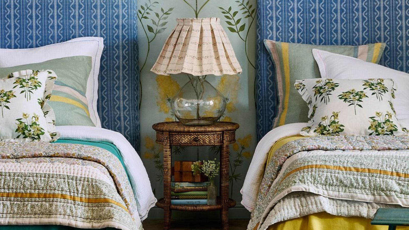 twin bedroom with matching blue patterned headboards and yellow printed bed linens with a nightstand in the middle with a glass bottom lamp and a pleated lampshade