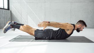 A man performs back extension exercises in an empty room on a yoga mat. He lies face down, with his legs straight behind him and arms straight at his sides; both his legs and arms are lifted off the floor.