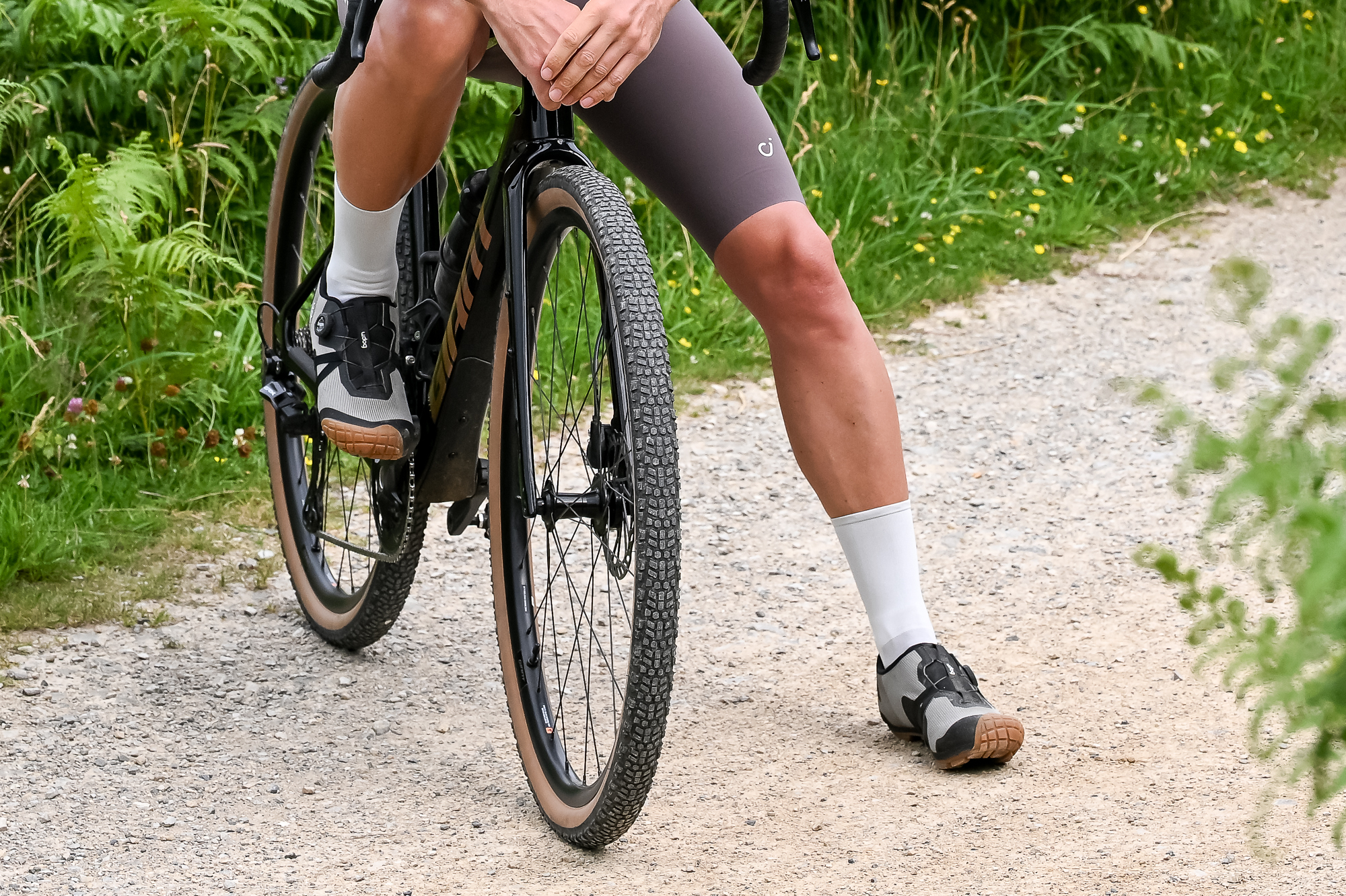 lower half of a man sitting on a bike wearing brown shorts, white socks and grey shoes on a gravel trail