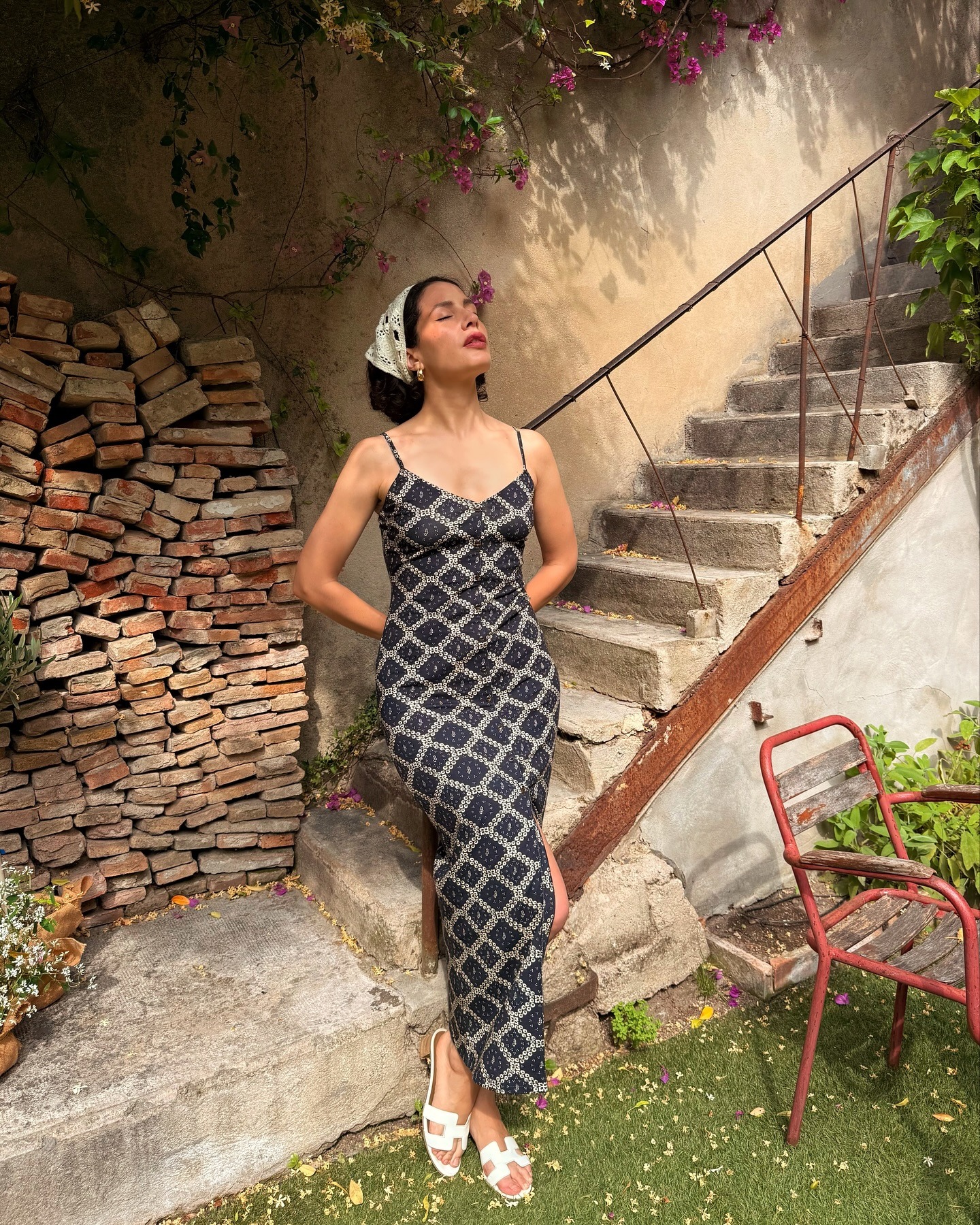 Influencer @melodiebanfield leans on the railing of a staircase outside, wearing a long-line printed slip dress with white sandals and a white bandana over her hair.