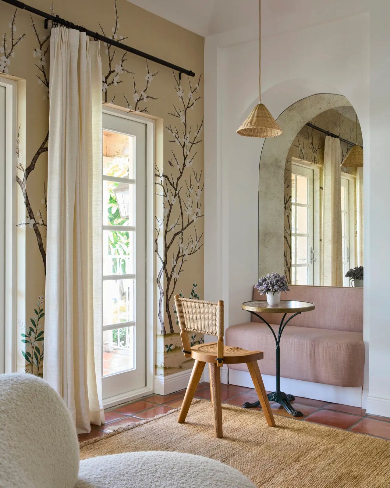 corner of room with floral cherry blossom wall mural on wall with white drapes, a small pink banquette seat in an arched niche with a mirrored wall, small table, rattan chair and pendant light hanging over head, with a jute rug on the floor
