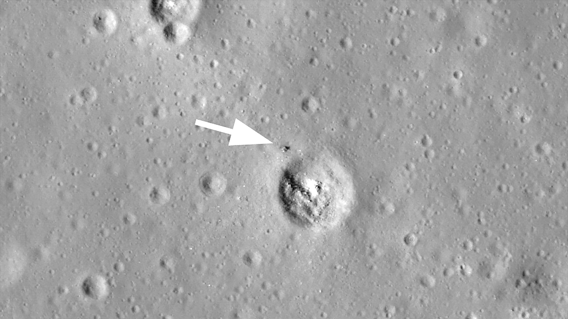 a black-and-white photo taken from the moon's orbit showing the location of where a robotic probe landed near a crater (with a superimposed white arrow pointing to the site).