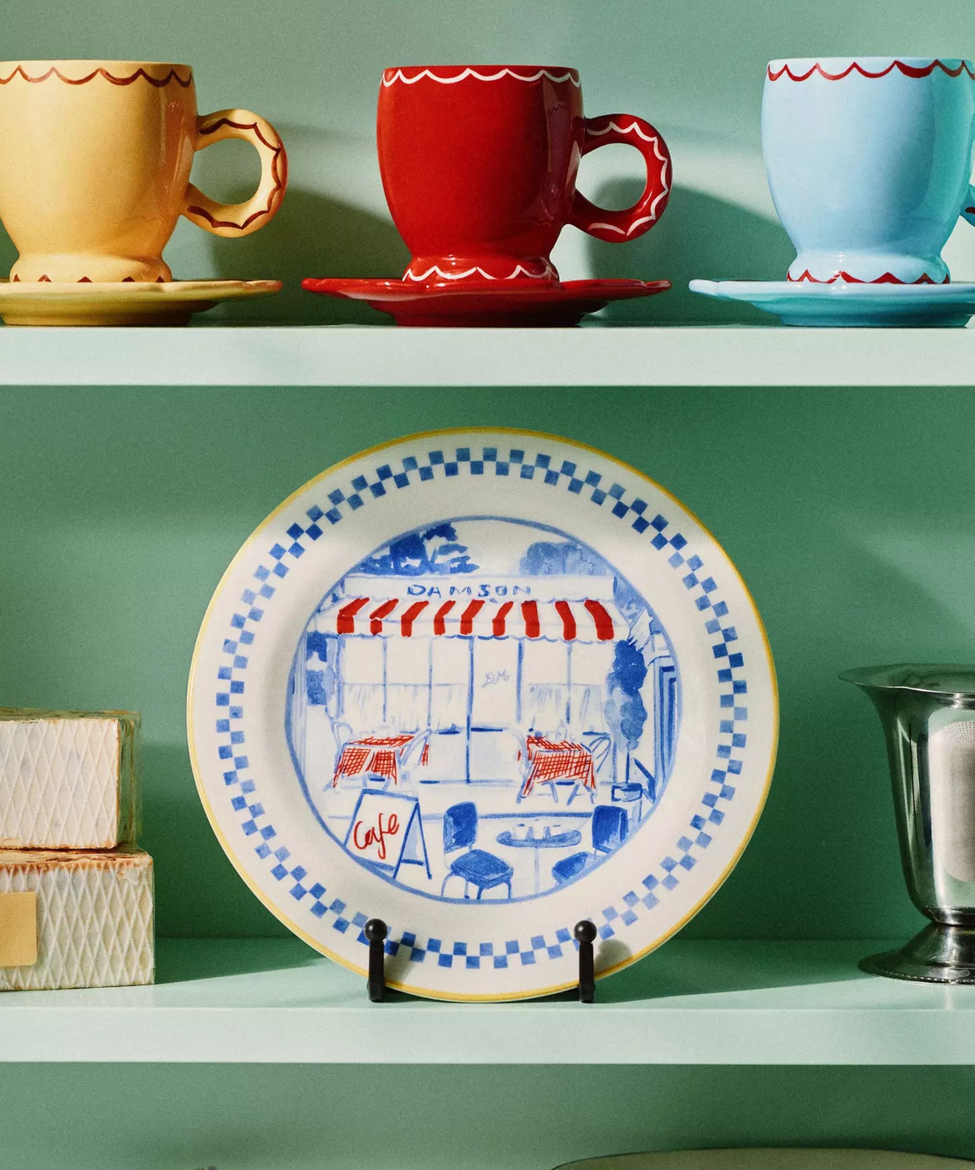 A green shelf displaying hand-painted plates and mugs from the Anthropologie x Damson Madder collection