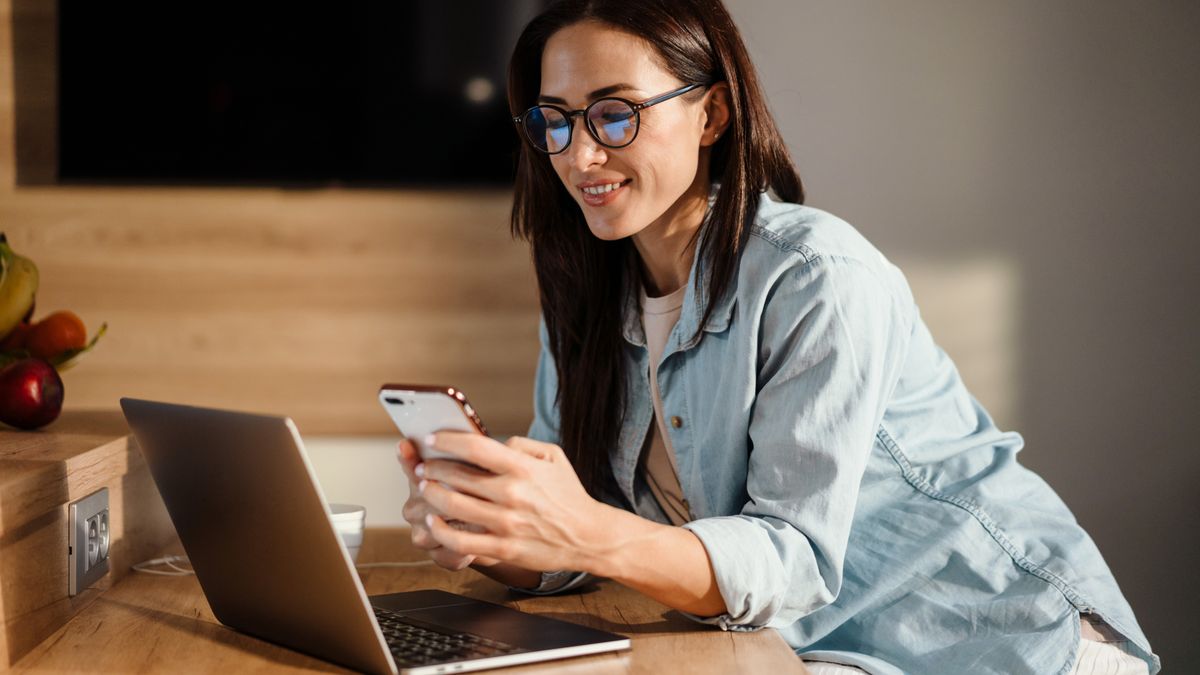 A woman sitting at a table and using a phone and a laptop