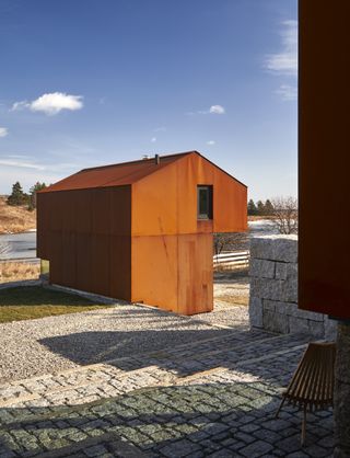 Close up exterior view of a Corten steel pavilion structure at Smith House under a blue, cloudy sky. The pavilion is surrounded by grass and stone flooring and there is brown grassy land and trees nearby