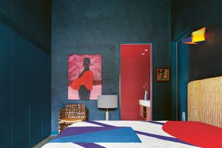 navy blue color drenched bedroom with colorful bed linen, artwork on wall with chair next to it, and view to bathroom with red walls