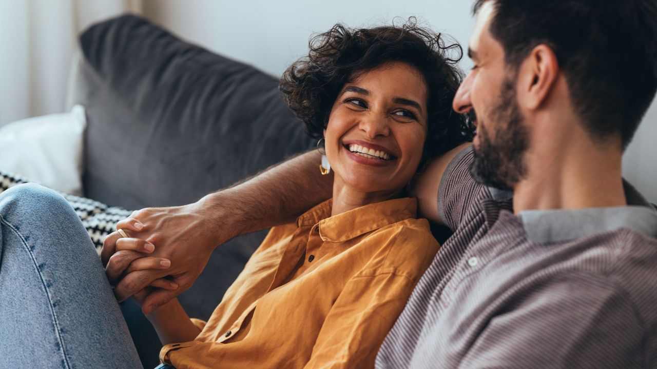 Man and woman sitting on the sofa together, woman laughing and smiling looking up at man, showing intimacy in a relationship