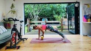 Woman on hands and toes on exercise mat, holding one kettlebell underneath her. She is in a domestic setting and wears white trainers, sports leggings with a floral print, and a pink vest top.