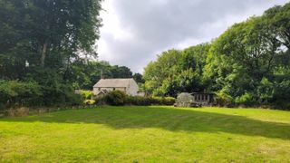 An acre of green land surrounded by trees with a stone cottage and greenhouse