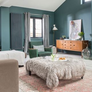 Teal painted living room with oversized footstool covered in faux fur throw and a vintage sideboard