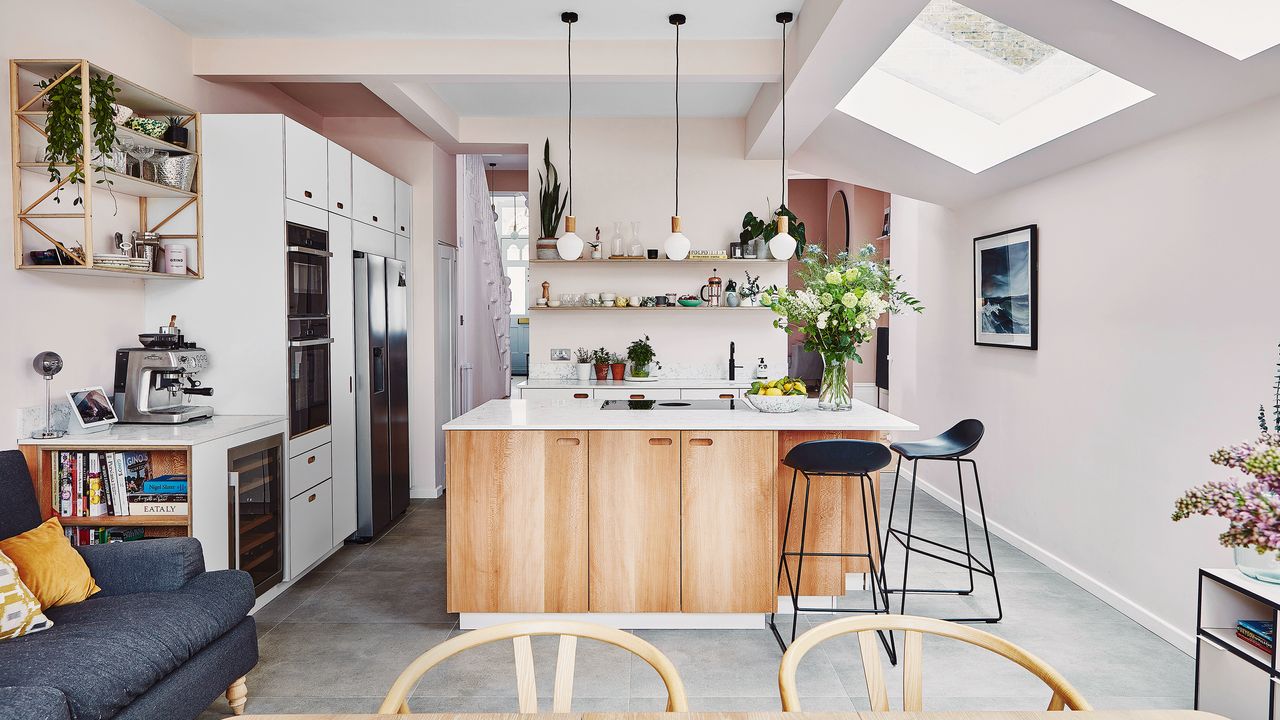 A modern kitchen-lounge with pink wall paint, trio of white pendant light fixtures, wooden island, built-in white cabinetry, oven appliance, coffee machine and wine fridge