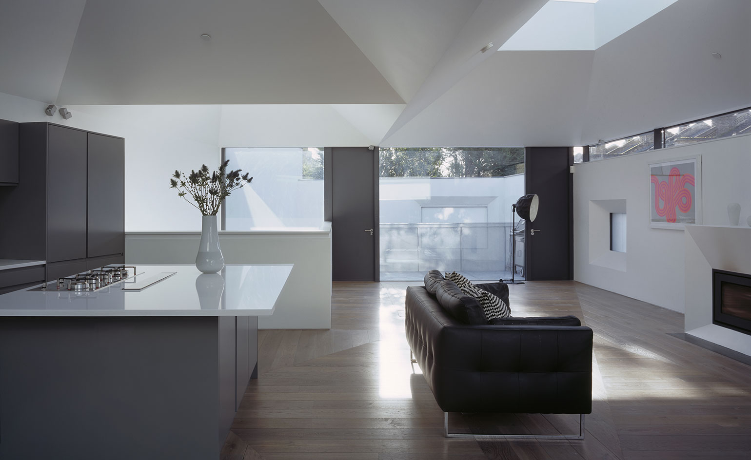 An open plan kitchen with wooden floors, gray cabinets, white ceramic top island (white vase with plant on the island) and a structural designed white ceiling. On the right of the kitchen is lounge area with dofa and a fire place