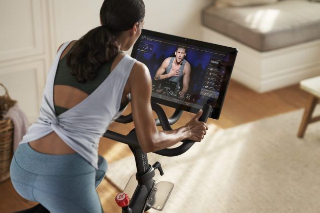 This image is of a woman with her back towards the camera, on a Peloton bike and looking at a instructor taking a class on a screen