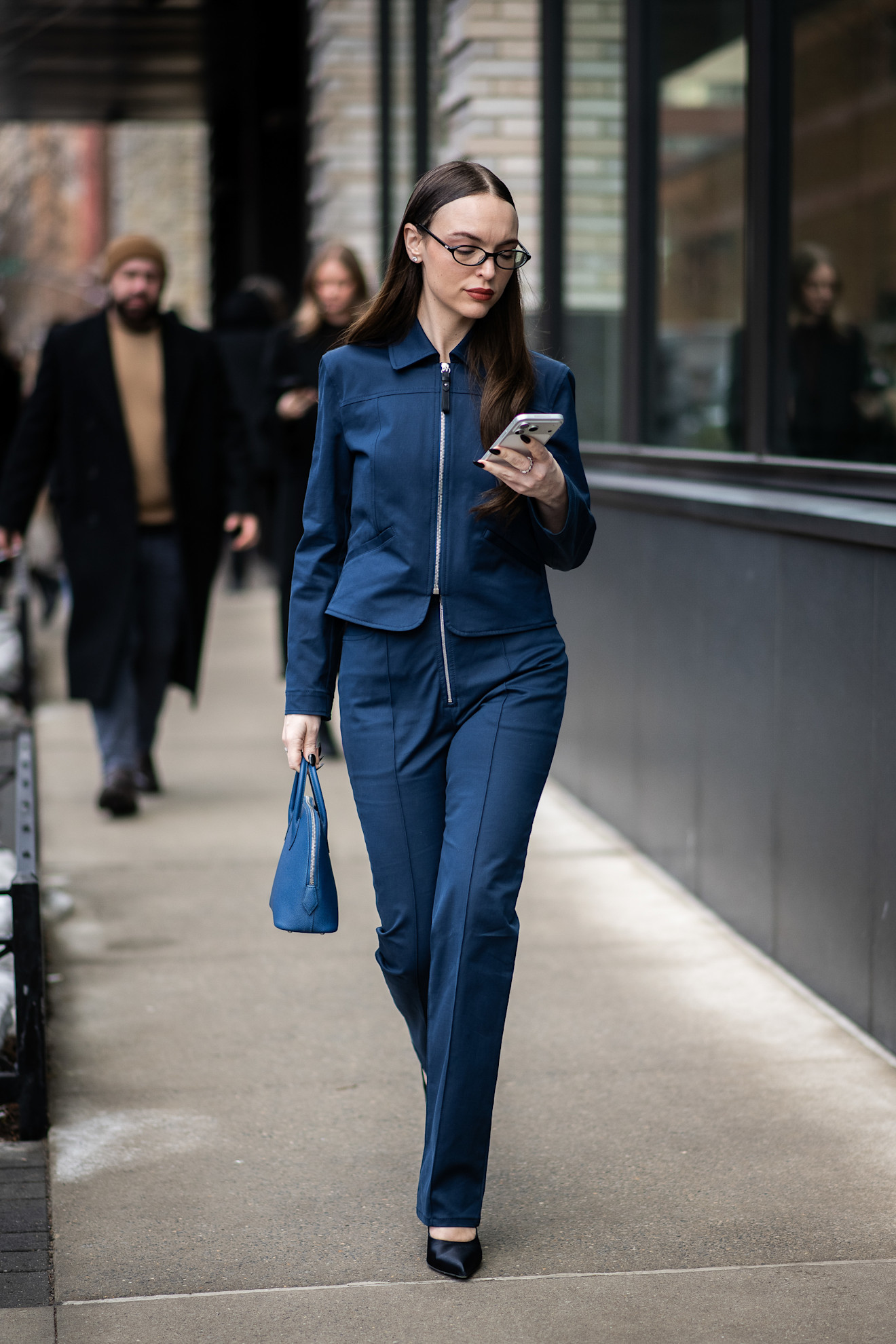 A fashion week attendee looking at her phone wearing blue jacket and pants matching set and eyeglasses