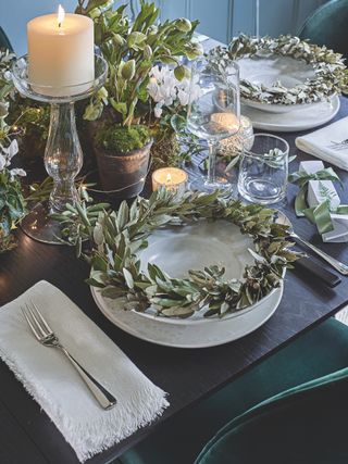 Christmas place setting with white candles, glass candlestick, silver green leaf wreaths and pots of greenery, ivy and moss