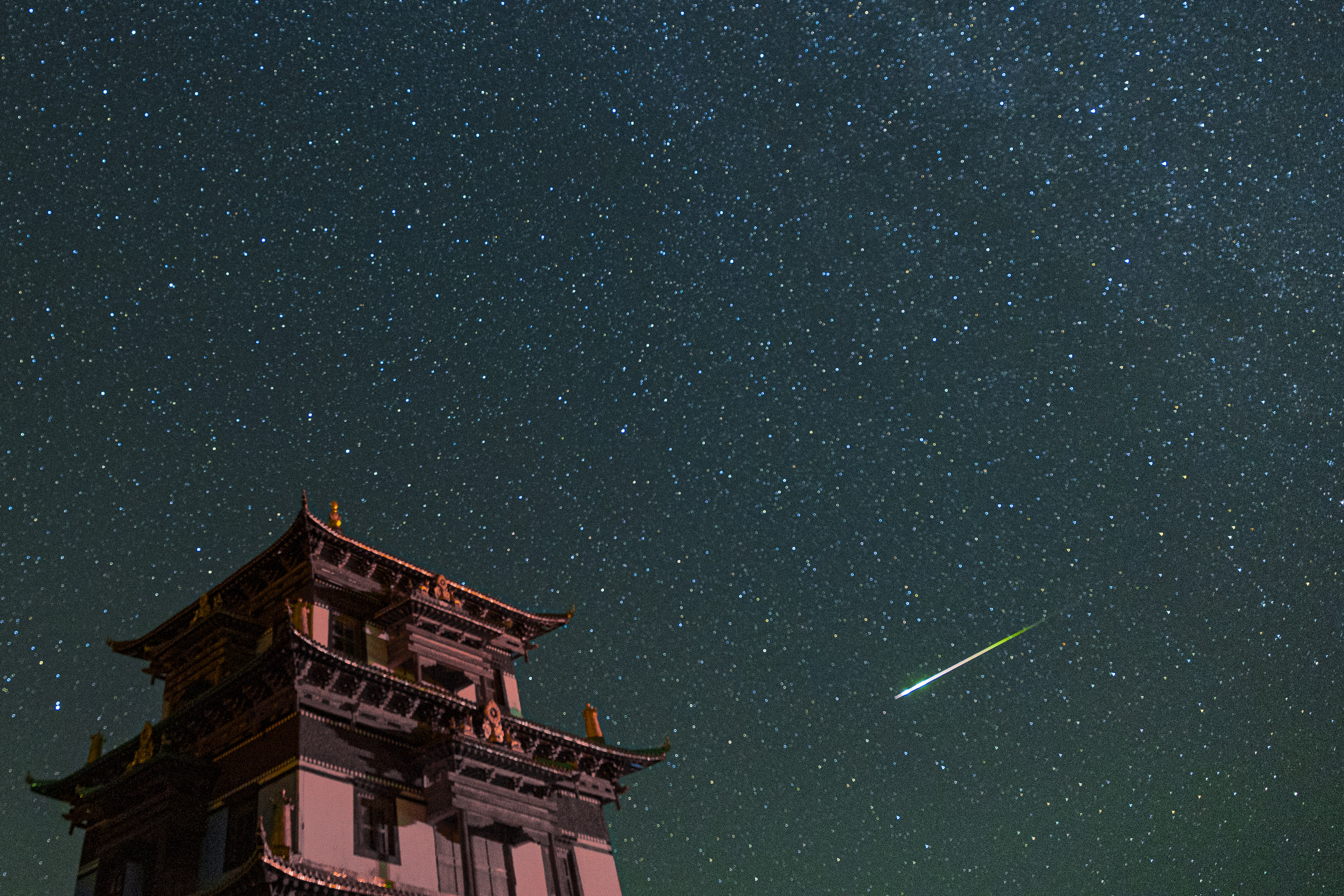 左側には大きな建物があり、満天の星空には明るい緑白色のペルセウス座流星が見えます。