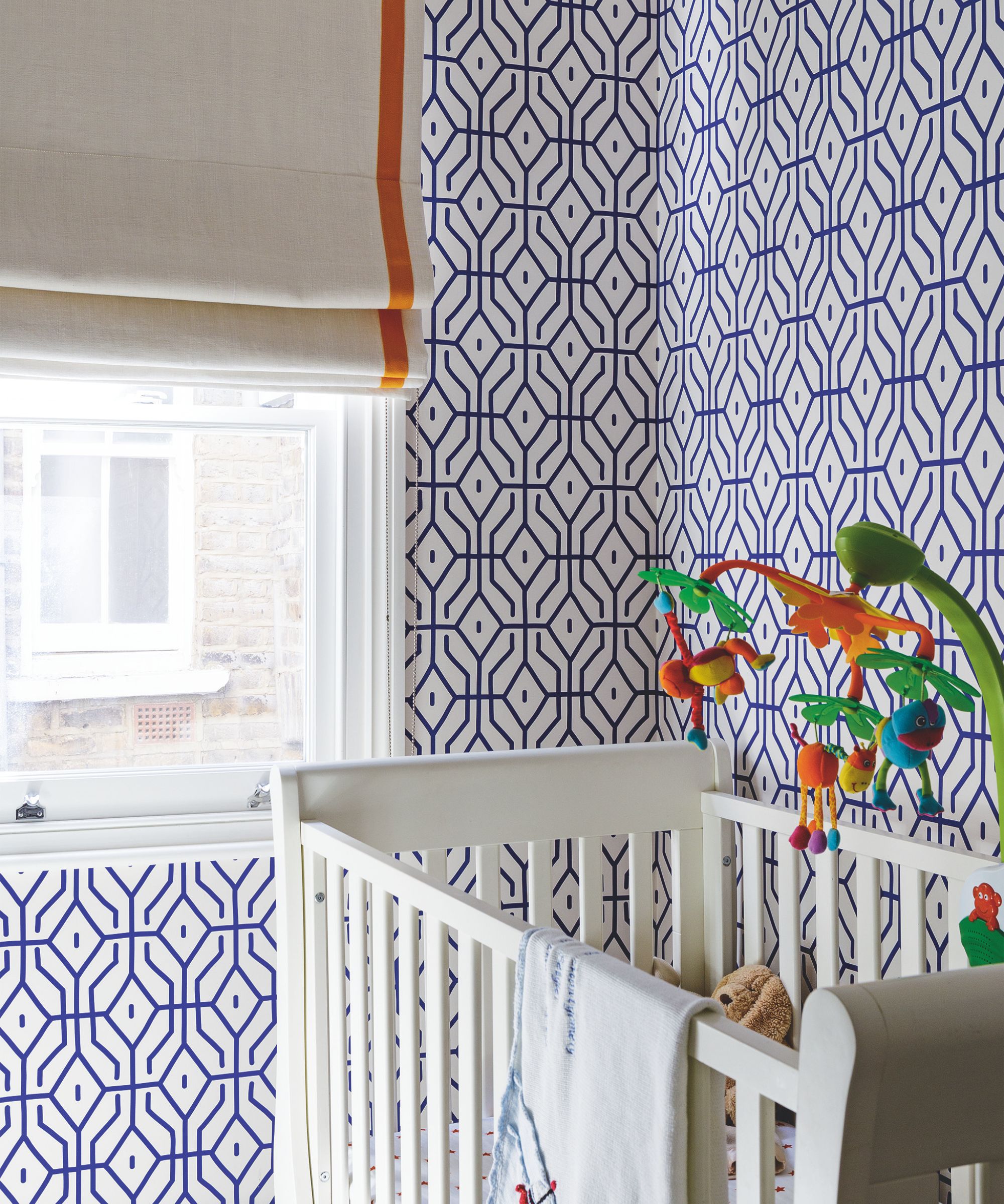 A nursery with white crib, blue and white geometric wallpaper, bright window and beige blind with orange stripe.