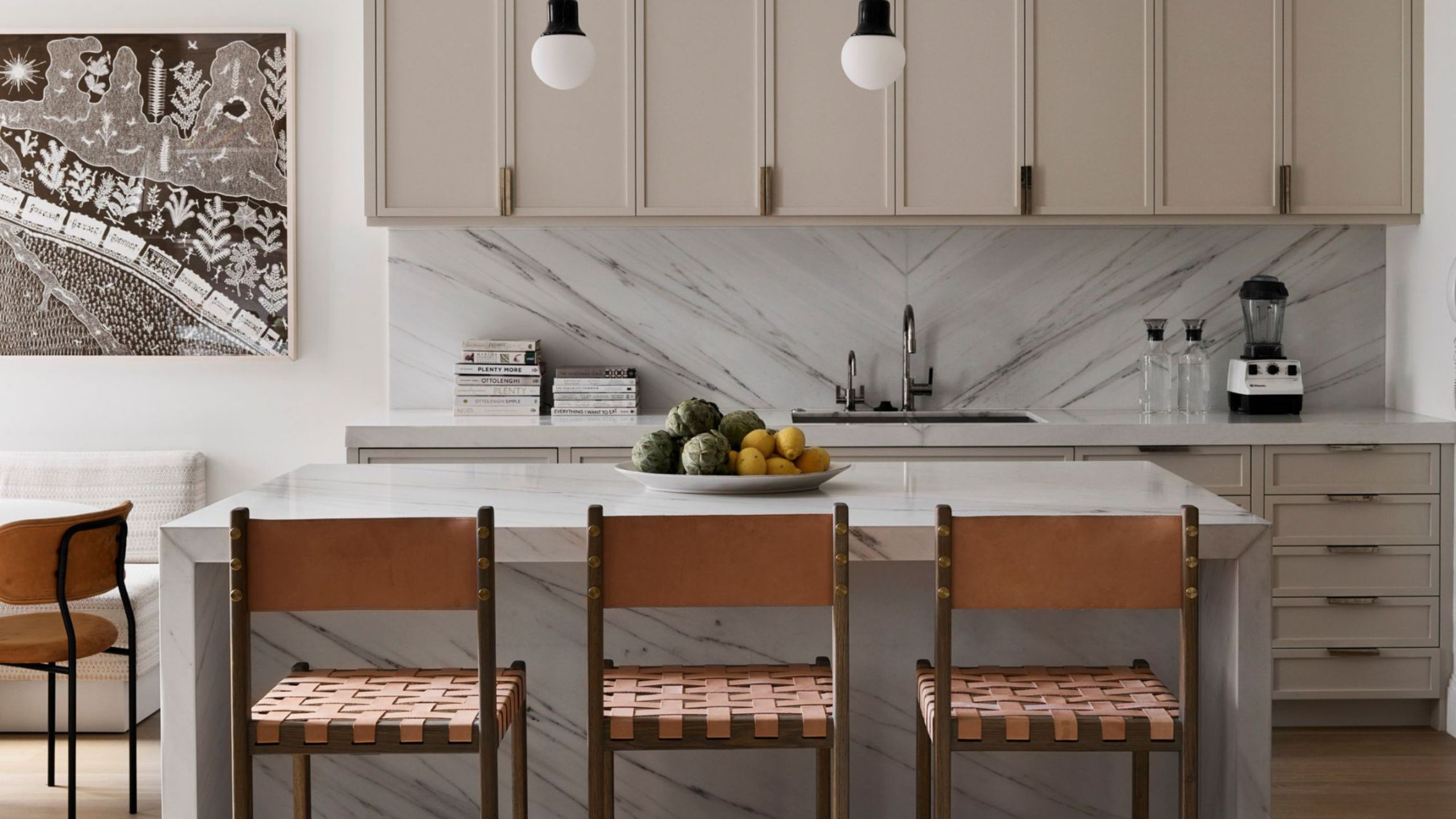 beige kitchen with marble island, beige cabinets, marble backsplash, stacks of books, a black-and-white artwork on the wall, and brown leather bar stools