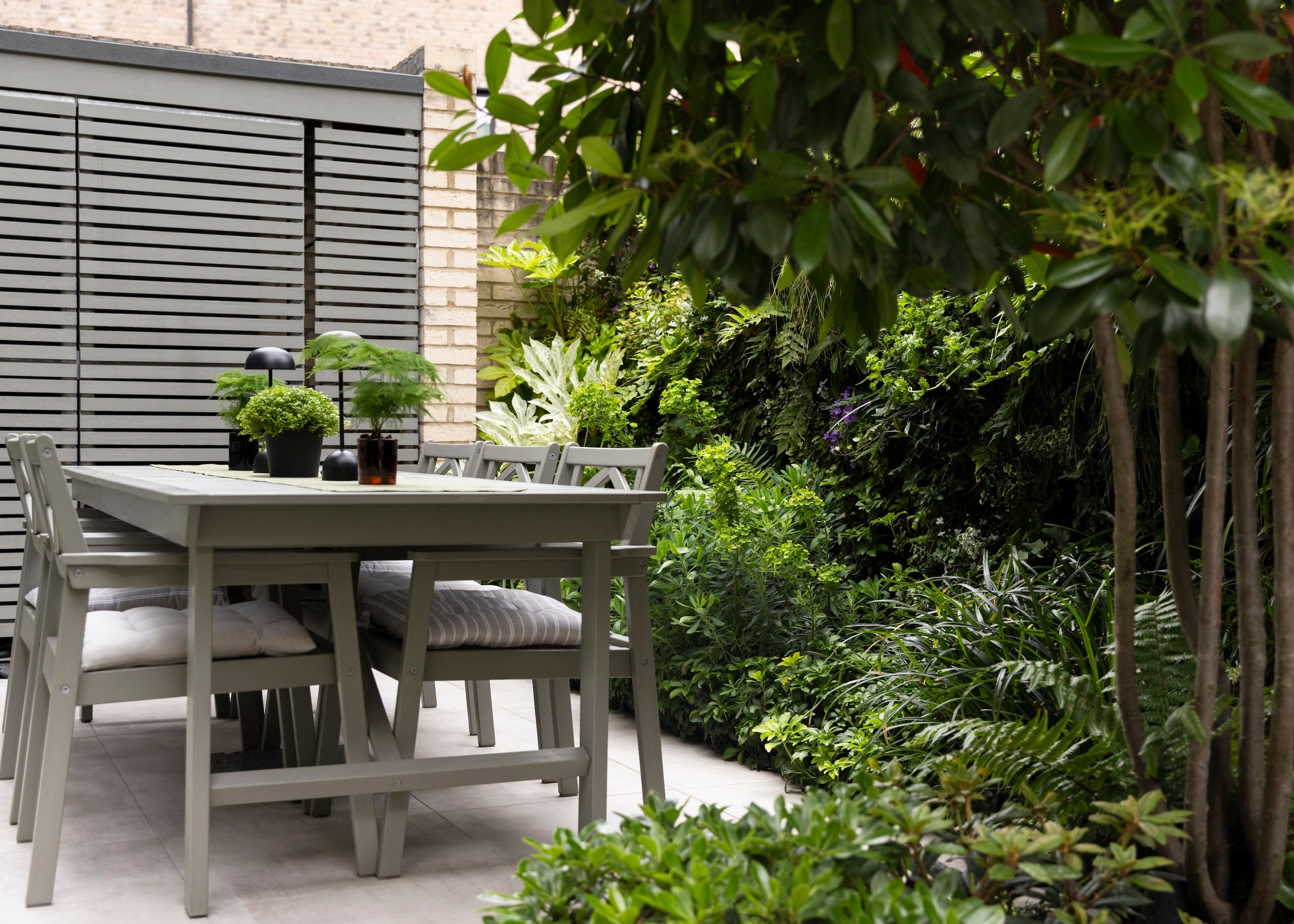 a grey dining table and chairs in the corner of a garden surrounded by green flower beds and vertical plantings