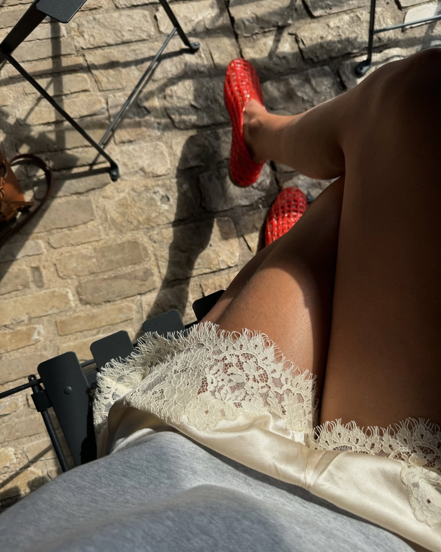 An overhead shot of style influencer Laura Vidrequin Roso sitting in an outdoor cafe chair wearing ivory Doen lace-trimmed silk satin shorts and The Row red jelly flats.