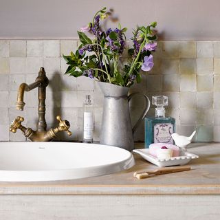 White bathroom basin showing tap, hand soap, silver vase with flowers and wooden toothbrush