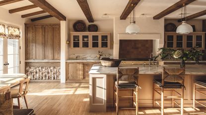 beamed neutral kitchen with wooden cabinets, island, wicker barstools and white pendant lights