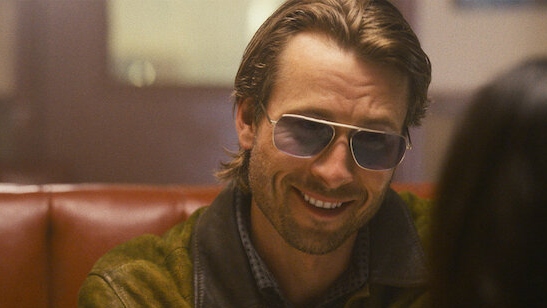 Glen Powell som Gary Johnson med solbriller og glat h&amp;aring;r, der smiler til Madison i en diner.
