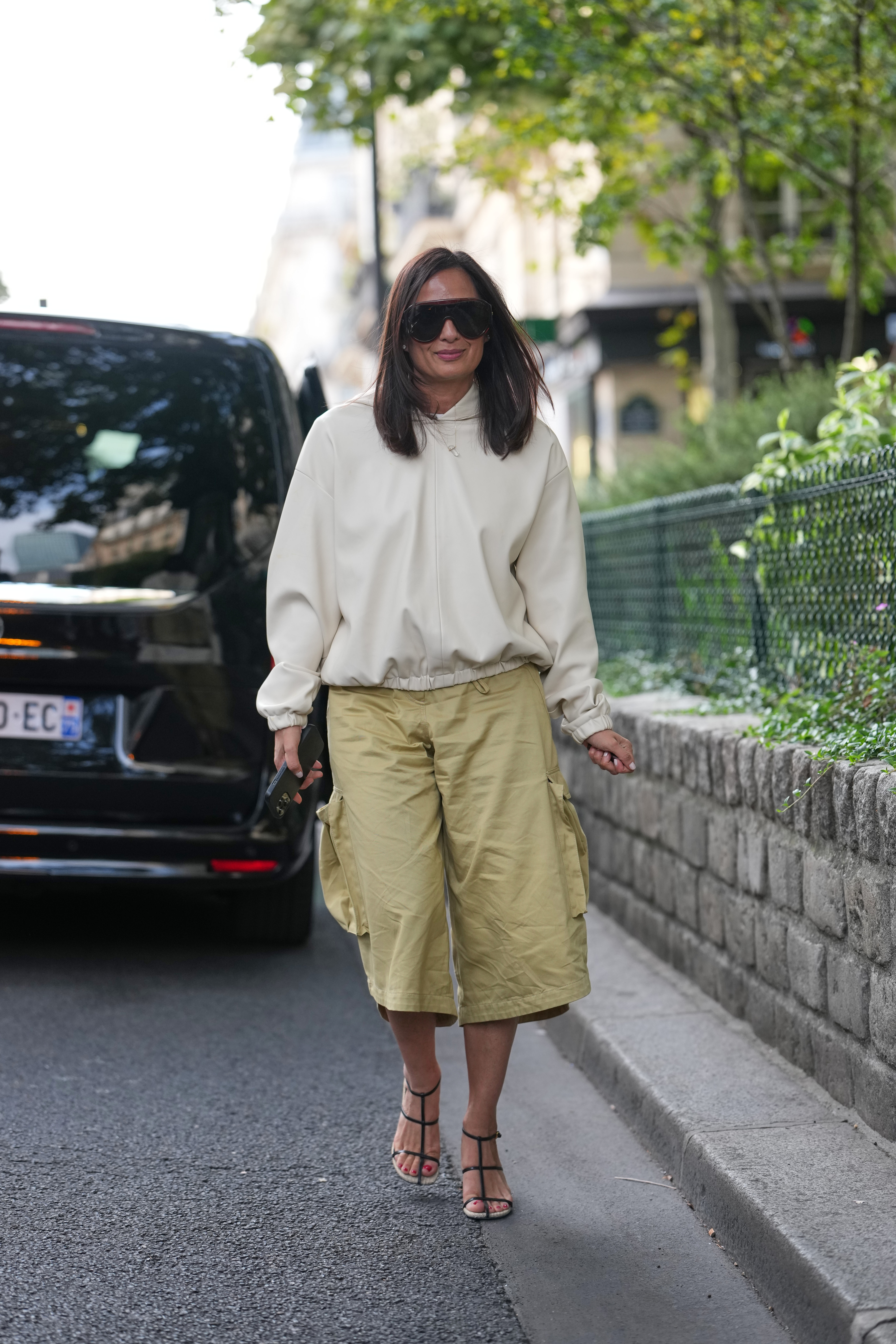 A guest wears an off-white pullover top, khaki cotton knee-length cargo shorts with large side pockets, black leather strappy heeled sandals on October 01, 2025 in Paris, France.