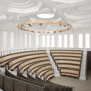 An amphitheater lecture hall with wooden seating, white walls and white ceiling. Light floods through the many tall windows that surround the upper part of the theatre.