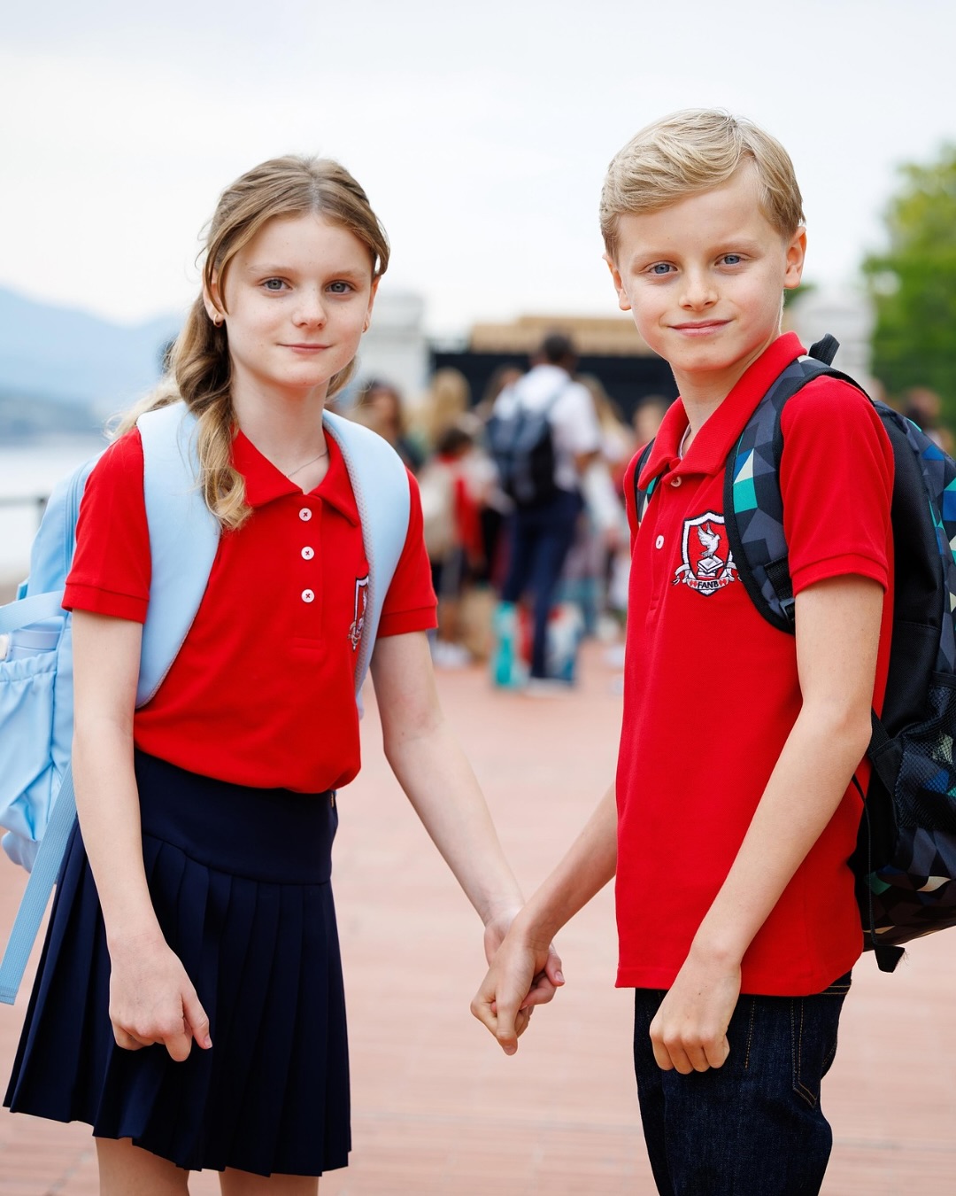 Prince Jacques and Princess Gabriella holding hands in red school polos wearing backpacks