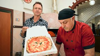 Phil Keoghan holding a pizza box and introducing the pizza challenge in Italy in Season 37 of The Amazing Race.