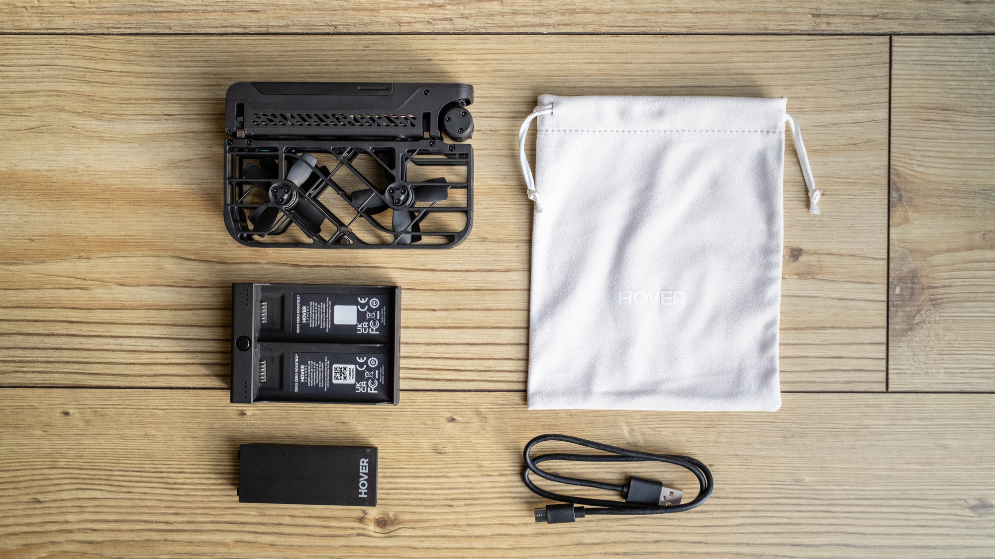 a folded black drone, batter charging block, charging cable, battery, and white carrying sack lay on a wooded surface.