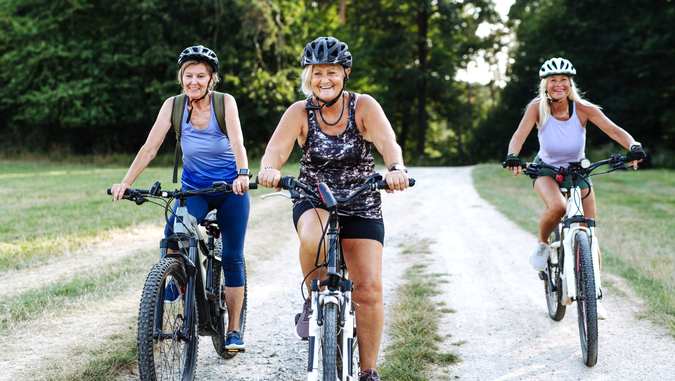 Three mature women riding e-bikes down a dirt trail.