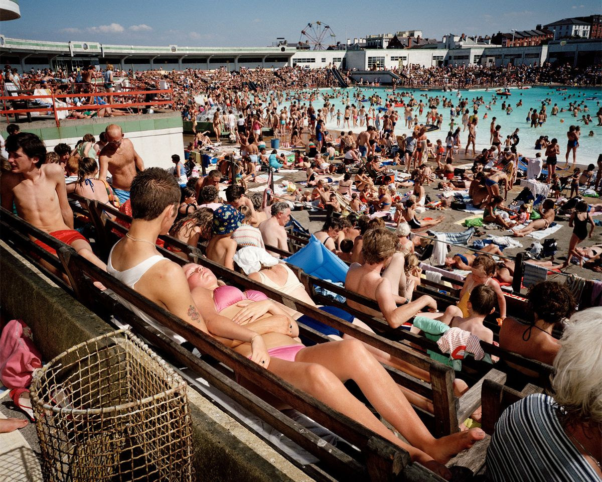 A densely crowded outdoor swimming pool area is filled with people sunbathing on wooden bleachers and swimming in a large turquoise pool under a clear blue sky.