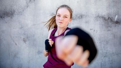 A young woman in workout clothes and light boxing gloves strikes a punch.