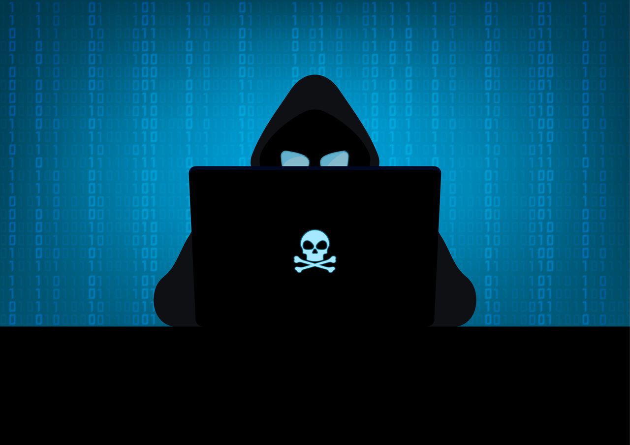 Silhouette of hacker wearing hood using laptop computer with glow in the dark blue skull and crossbones logo in dark room on blue binary code number background