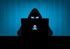 Silhouette of hacker wearing hood using laptop computer with glow in the dark blue skull and crossbones logo in dark room on blue binary code number background