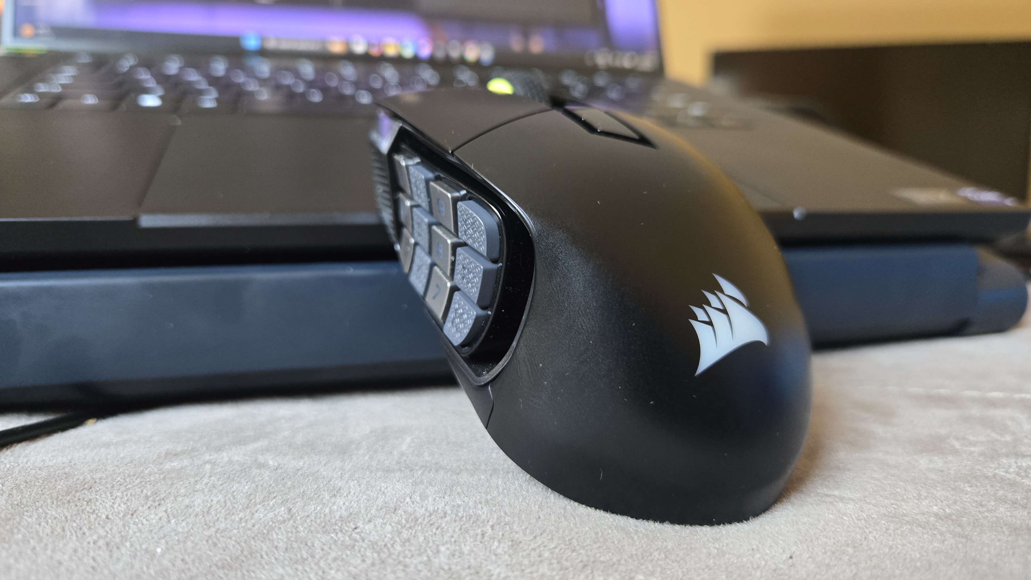 A Corsair Scimitar Elite Wireless SE gaming mouse on a carpet next to a gaming laptop and Stream Deck.