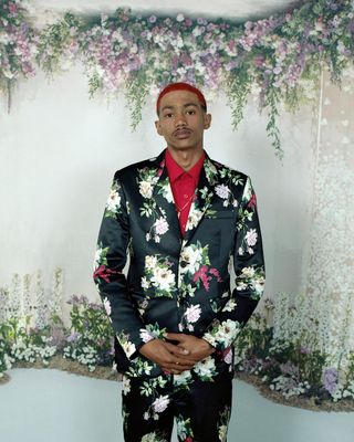 African man with dyed red hair wearing a floral black suite standing in front of floral patterned white curtain.